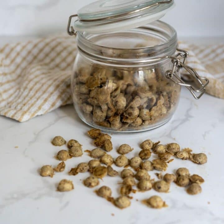 A glass jar with a metal clasp is filled with small, round, brown chicken sardine cat treats, some of which are scattered on a white worktop next to a striped cloth.