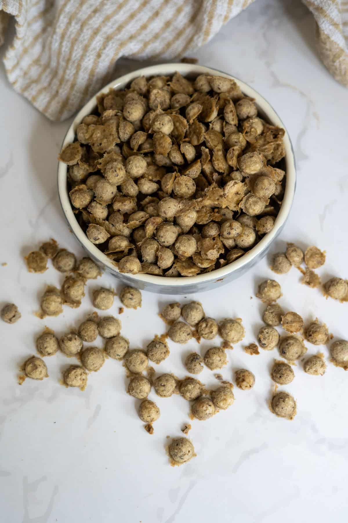 A bowl filled with round, brown cereal pieces—resembling chicken sardine cat treats—rests on a white marble surface, with some pieces scattered beside a beige striped cloth.