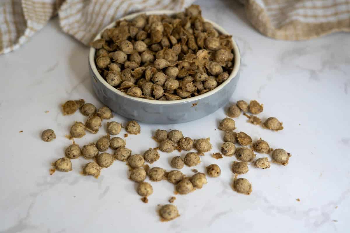 A bowl filled with small, round, brown chicken sardine cat treats, with some scattered on a white marble surface. A beige and white cloth is in the background.