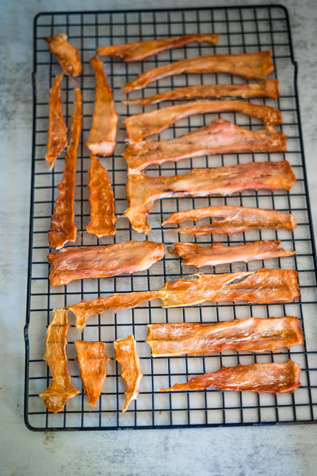 Thin strips of cooked jerky, perfect for low-fat dog treat recipes, are arranged on a black cooling rack set on a light-coloured surface.