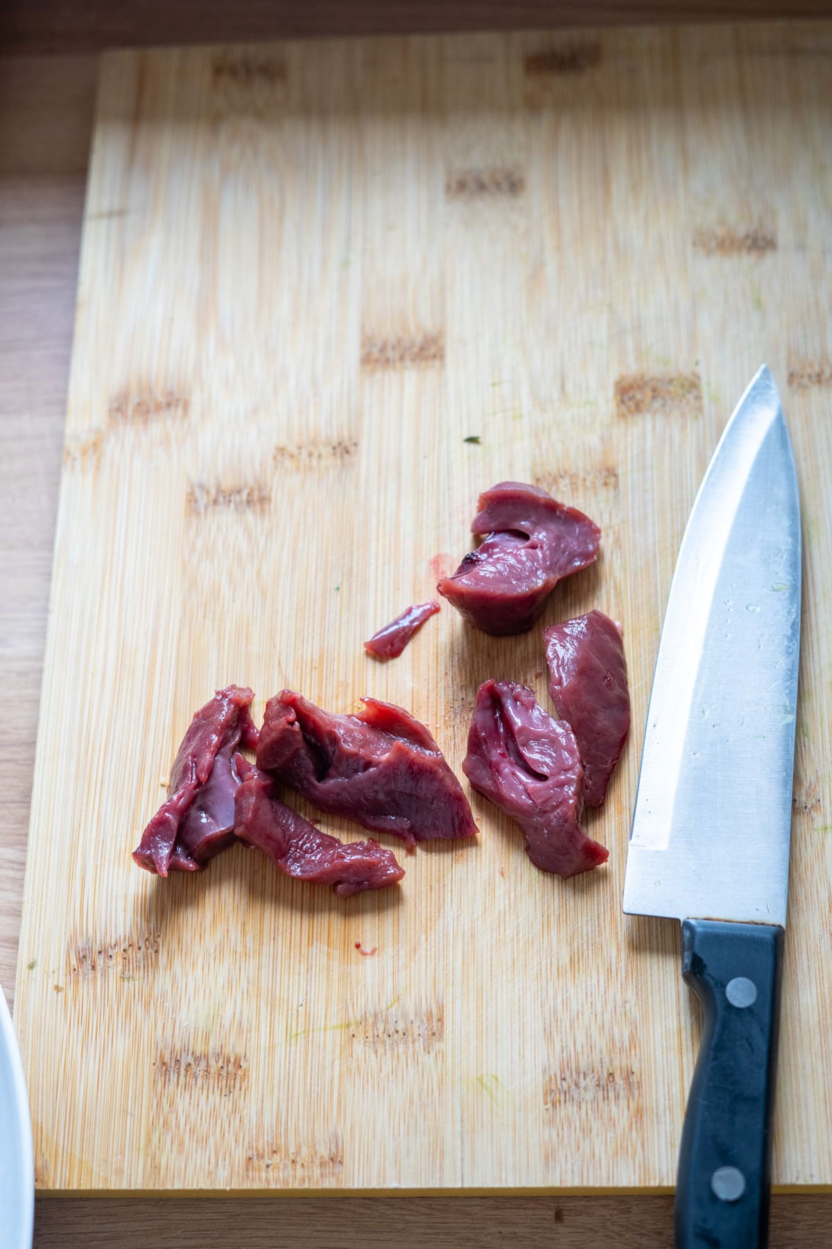 Sliced raw meat pieces on a wooden chopping board next to a large kitchen knife.