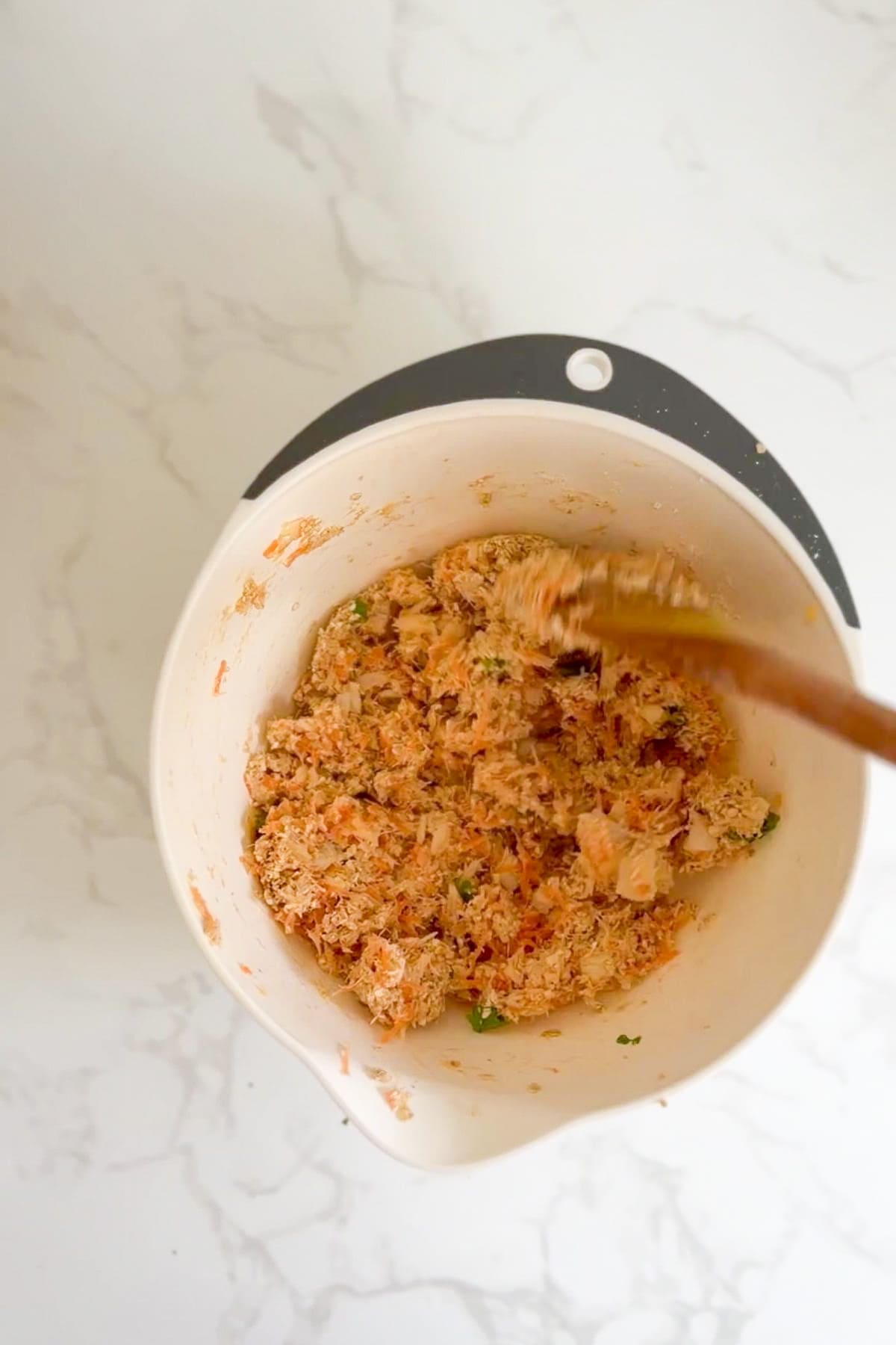 A mixing bowl containing a blended mixture of shredded cod and carrot for homemade dog treats, being stirred with a wooden spoon on a white marble surface.