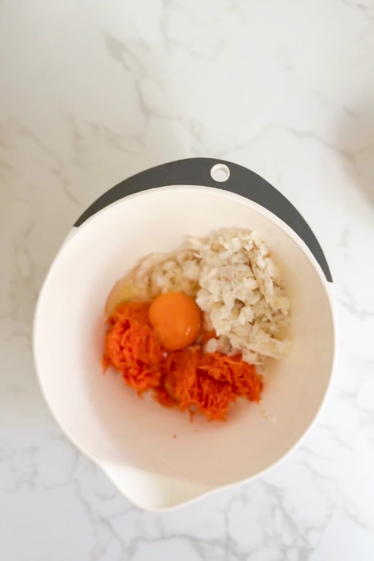 A mixing bowl containing a raw egg, grated carrots, and chopped onions sits on a marble worktop—perfect for preparing homemade cod carrot dog treats.