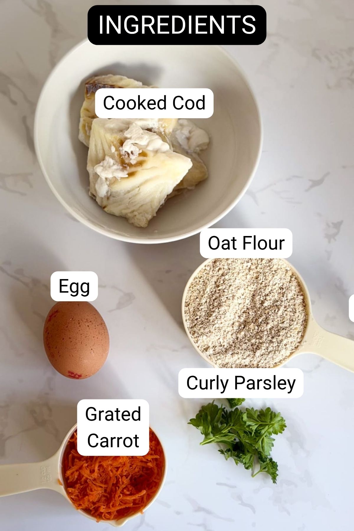 A bowl of cooked cod, oat flour in a measuring jug, an egg, grated carrot, and curly parsley arranged on a white surface—perfect ingredients for homemade cod carrot dog treats, each labelled with text.