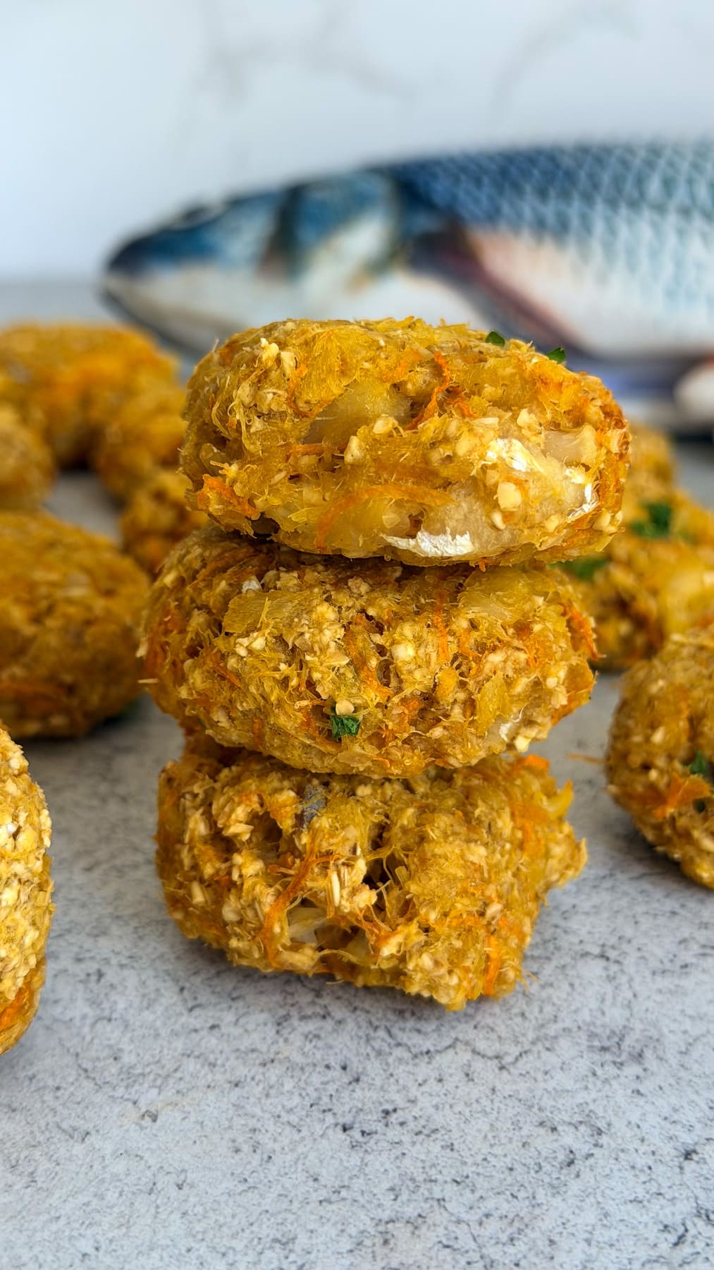 Three homemade fishcakes stacked on a grey surface, with extra fishcakes and a whole raw cod in the background—perfect inspiration for making nutritious cod carrot dog treats at home.