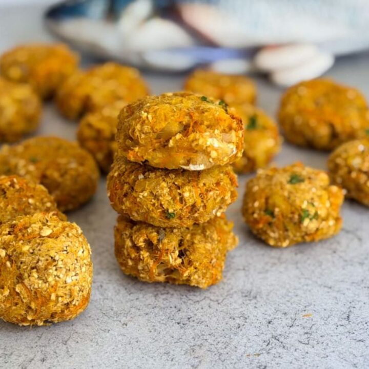 A stack of three golden brown fishcakes sits on a grey surface, with several more fishcakes scattered around in the background.