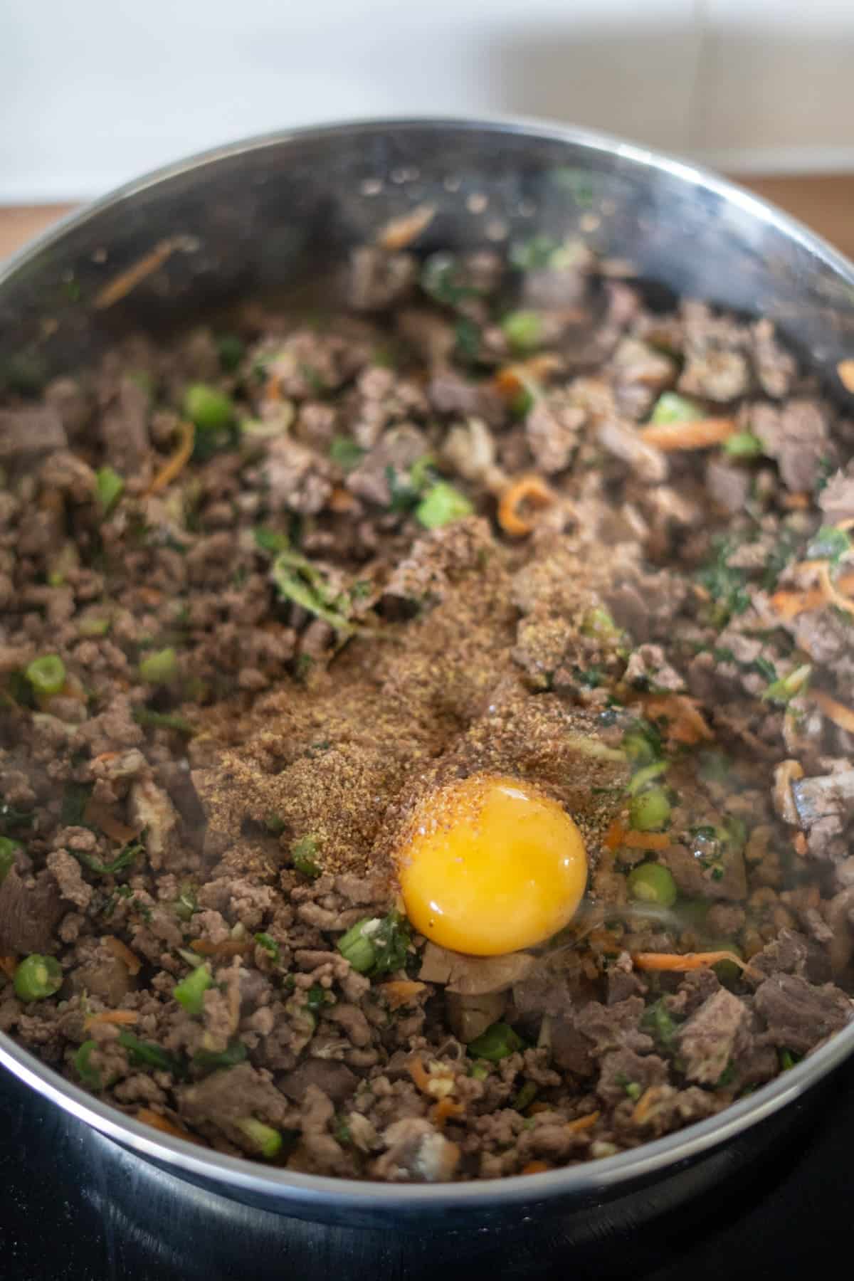 Minced meat, chopped vegetables, and seasonings are being cooked in a pan, with a raw egg cracked on top in the centre—perfect for trying out a homemade beef dog food recipe.