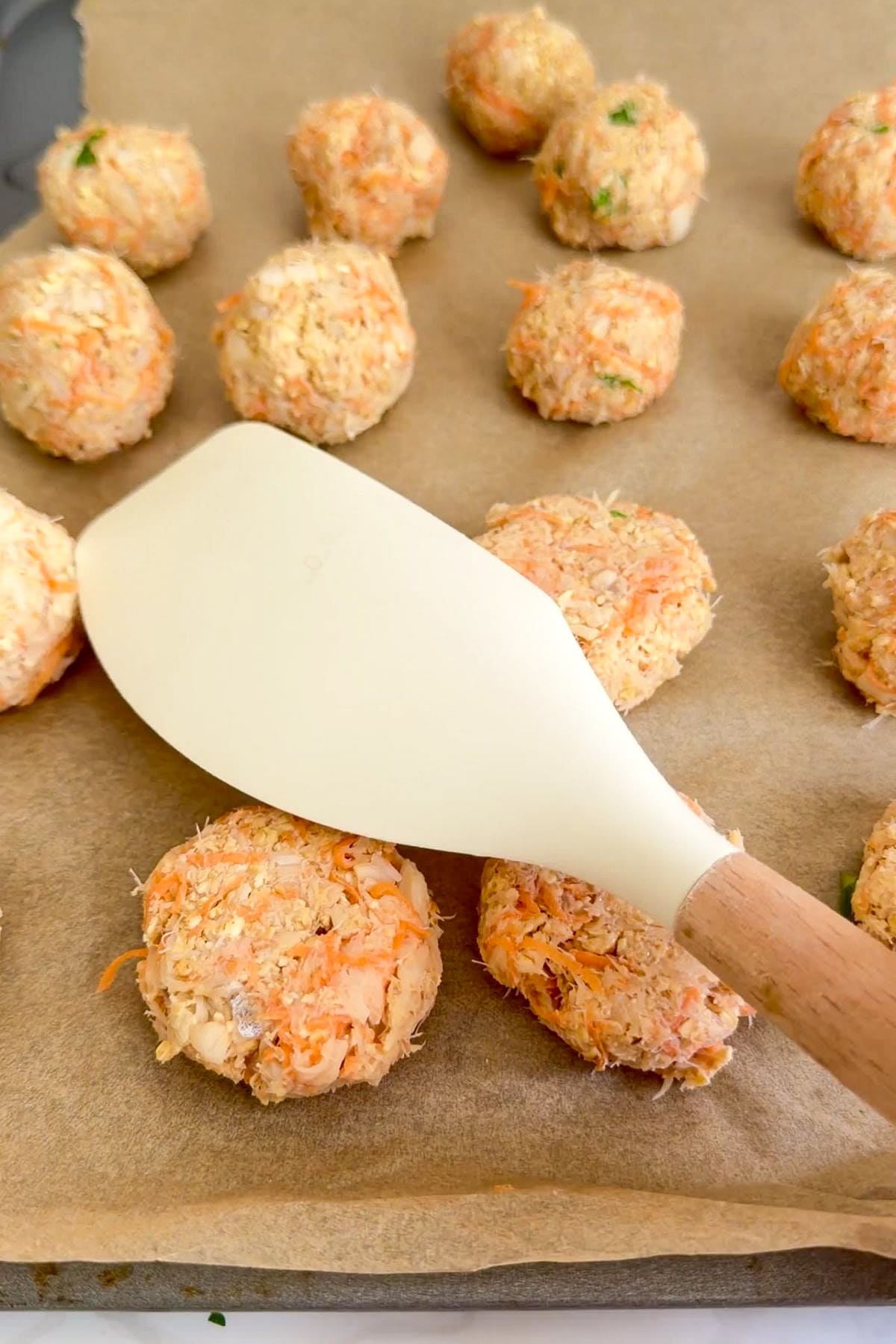 A white fish slice presses down on a round, uncooked salmon mixture ball for cod carrot dog treats on baking parchment, with more salmon balls in the background.