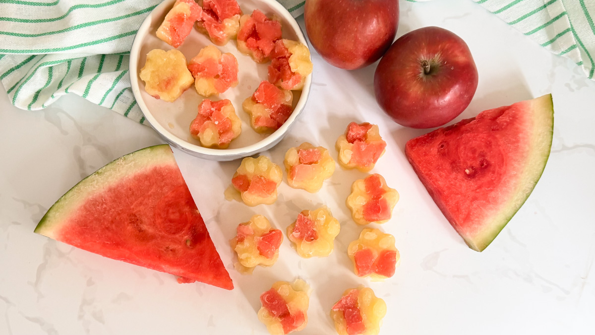 Slices of watermelon and apples are arranged with small paw-shaped fruit sweets on a white surface next to a green-striped cloth.