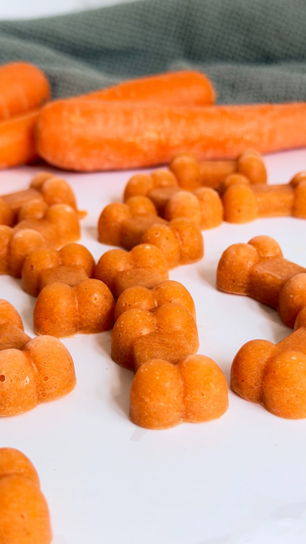 Orange, paw-shaped carrot bone broth dog treats are arranged on a white surface with whole carrots and a green textured cloth in the background.