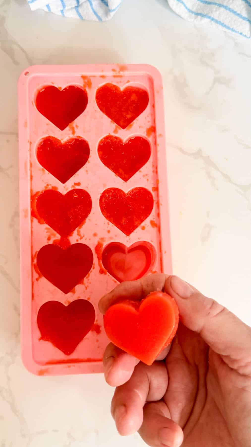 A hand holding a red heart-shaped sweet above a pink silicone mould filled with similar heart-shaped sweets on a white surface.