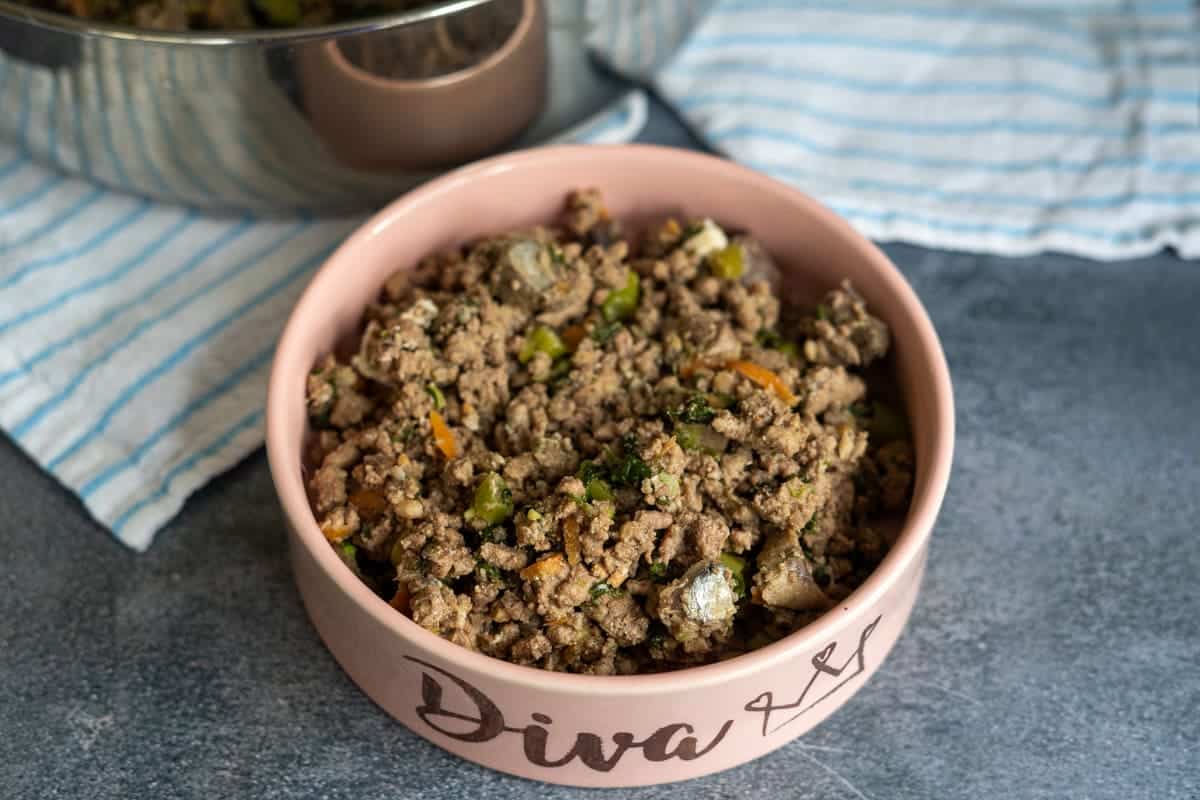 A pink pet bowl labelled "Diva" is filled with a beef dog food recipe of cooked minced meat mixed with vegetables, placed on a grey surface next to a striped cloth.