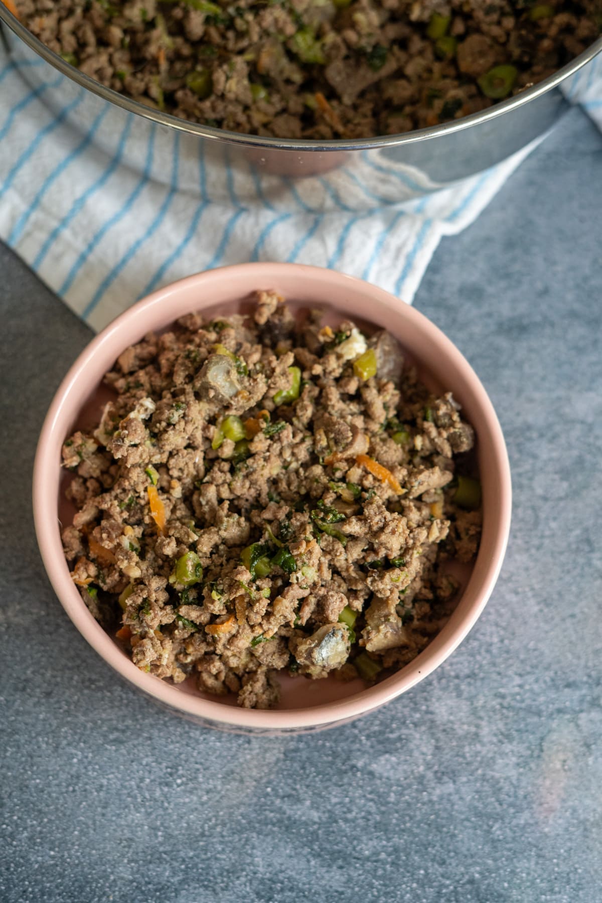 A bowl of cooked minced meat mixed with vegetables, prepared as a homemade beef dog food recipe, sits on a grey surface next to a pot and a striped cloth.