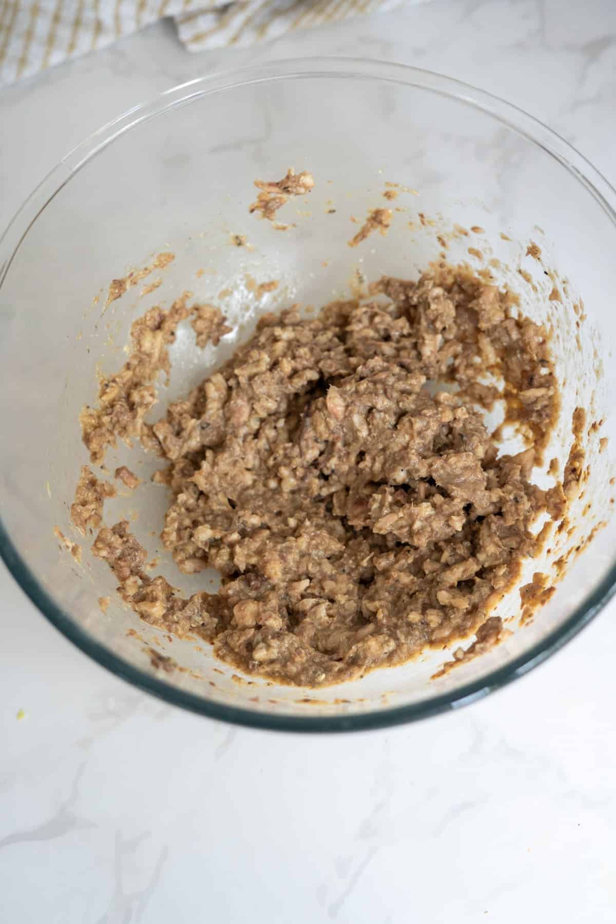 A glass mixing bowl containing a thick, chunky brown mixture for homemade chicken sardine cat treats sits on a white marble worktop.