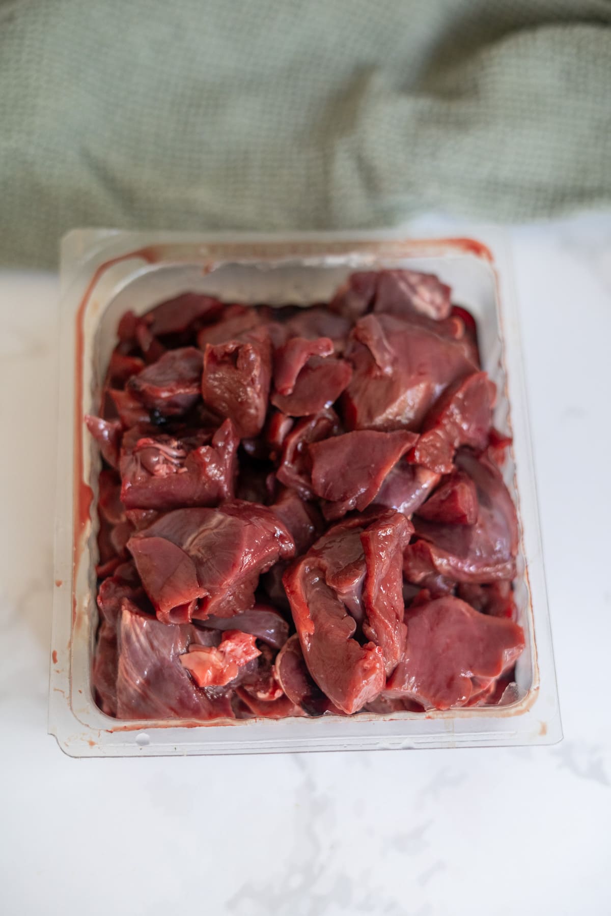 A plastic container filled with raw, chopped pieces of offal on a white surface with a green cloth in the background.