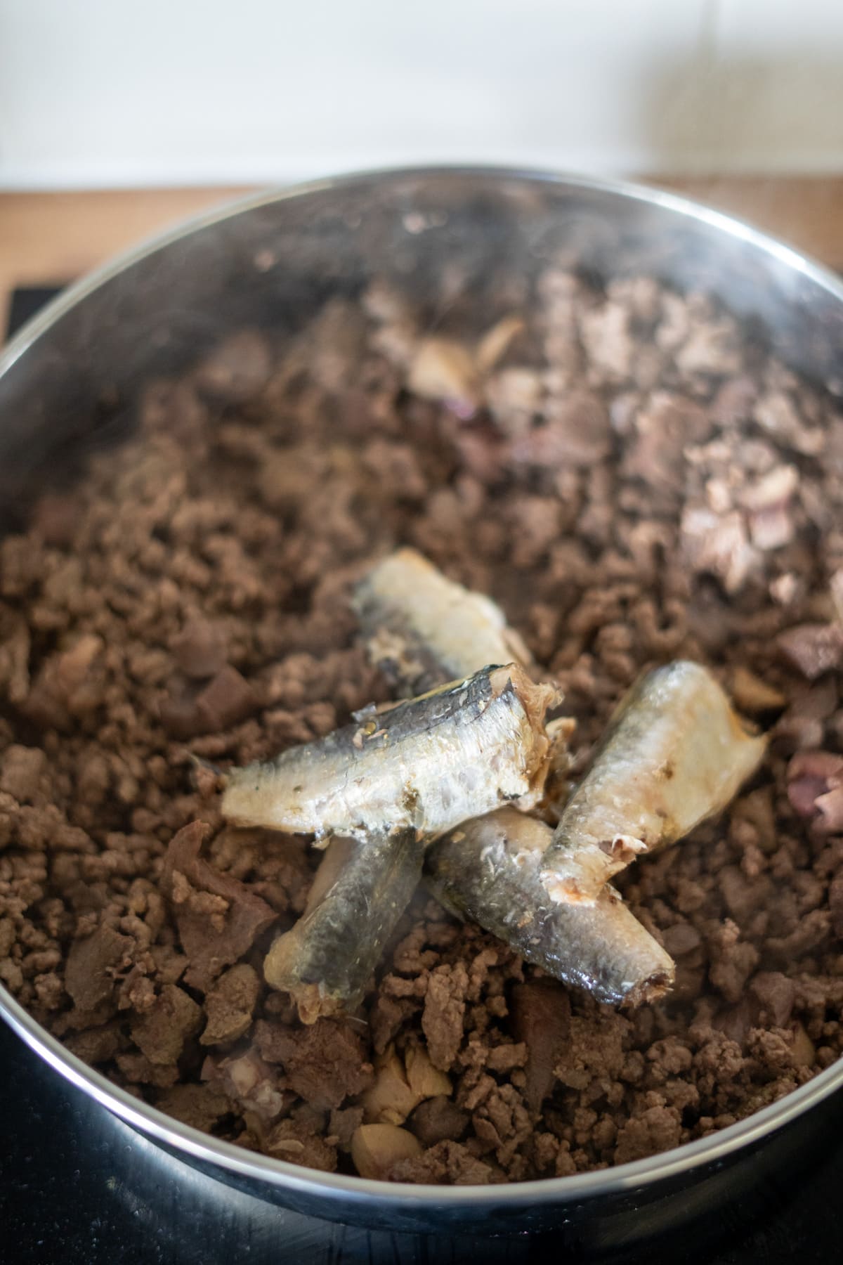 A close-up of a pan filled with cooked minced meat, perfect for a homemade beef dog food recipe, and several pieces of tinned sardines on top.