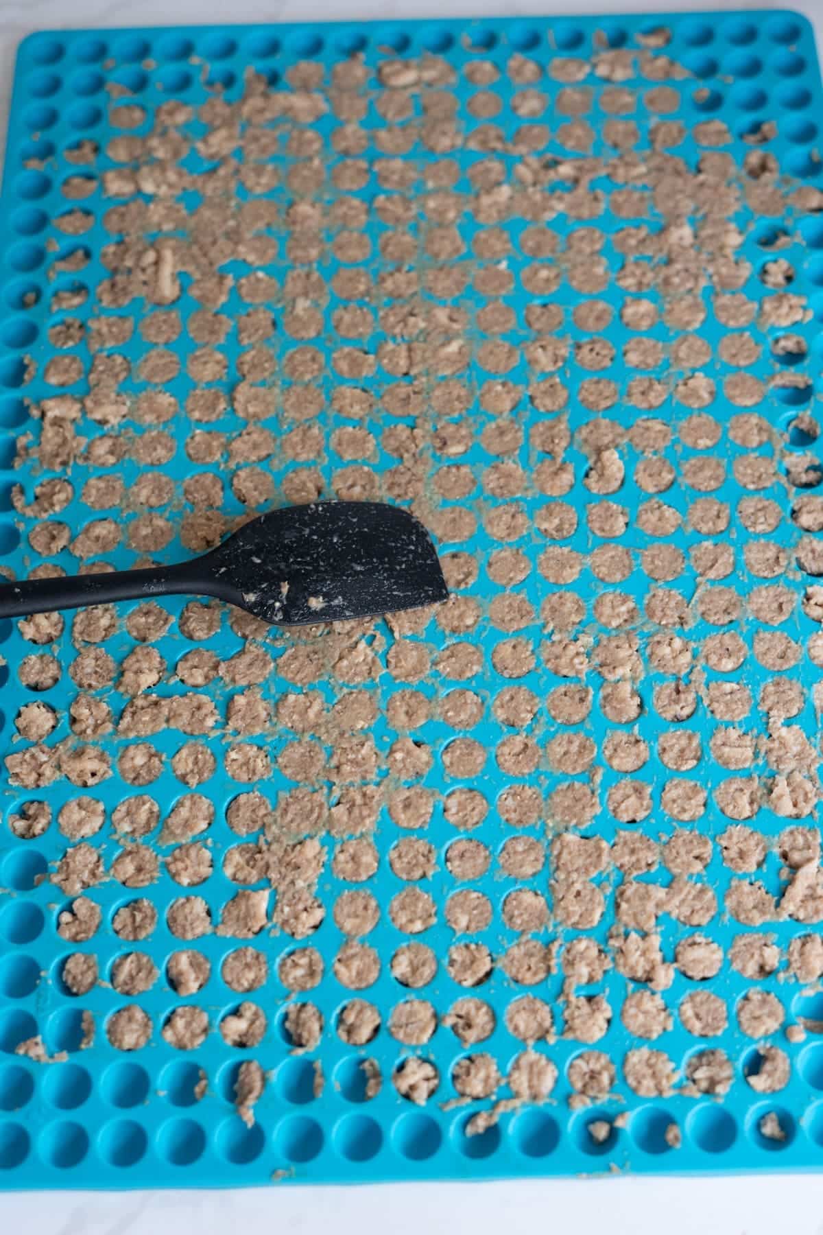A blue silicone mould filled with small round portions of a brown chicken sardine cat treats mixture, with a black spatula resting on top.