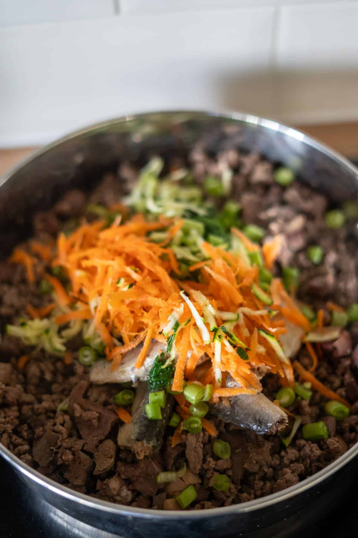 A pan filled with cooked minced beef, topped with grated carrots, grated courgette, chopped green beans, and a few small fish—perfect as a wholesome beef dog food recipe.