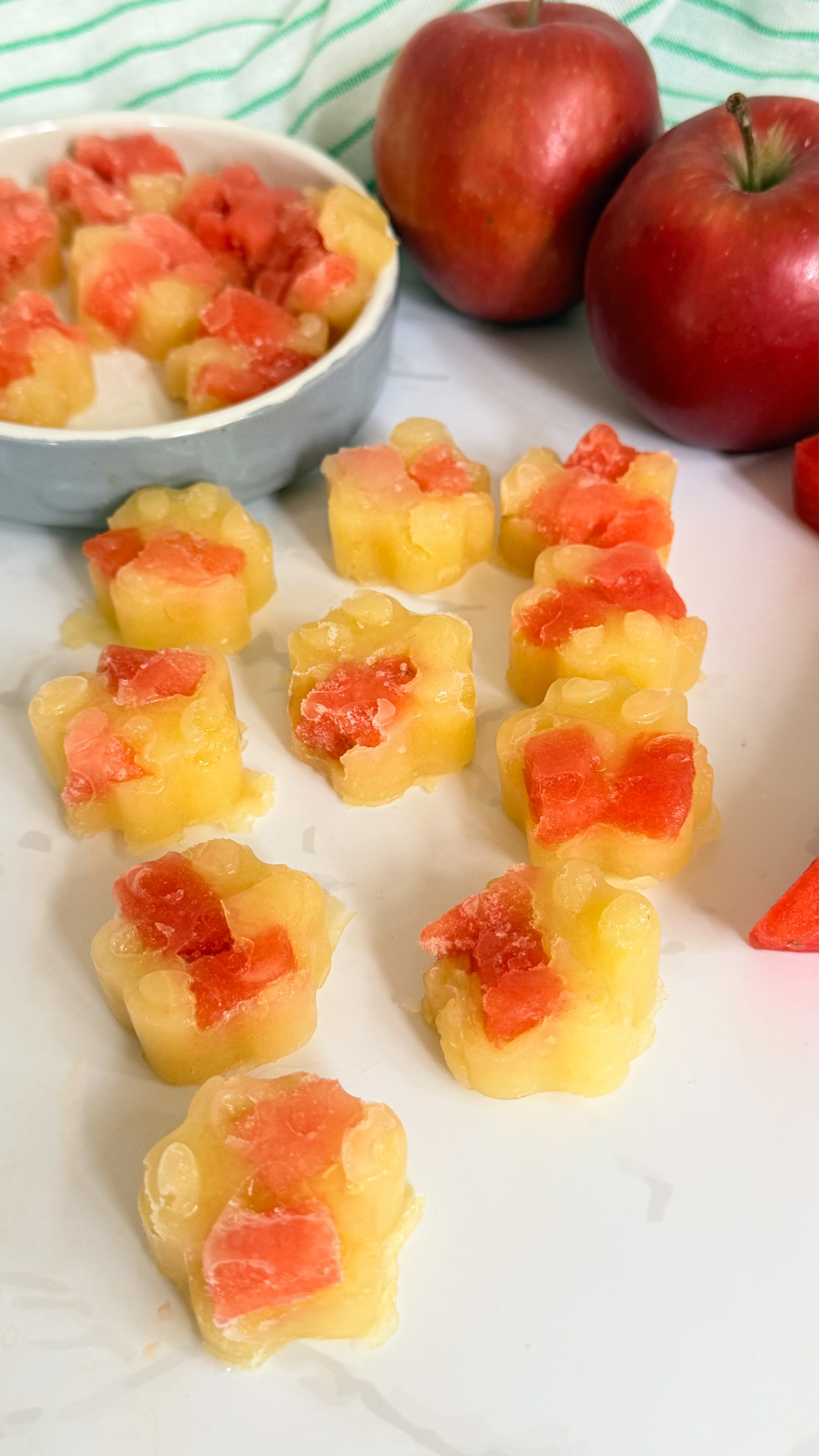 Small, flower-shaped frozen treats with red and yellow sections are arranged on a white surface, with apples and more treats in the background.