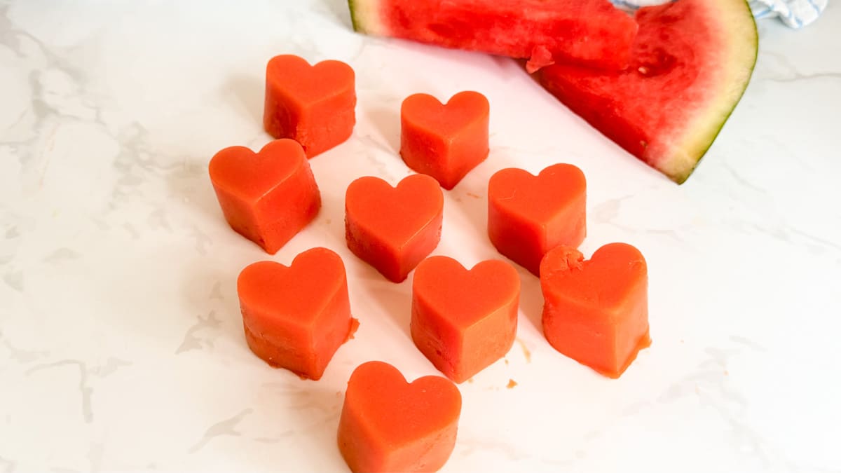Nine heart-shaped watermelon pieces are arranged on a white surface, with a watermelon slice in the background.