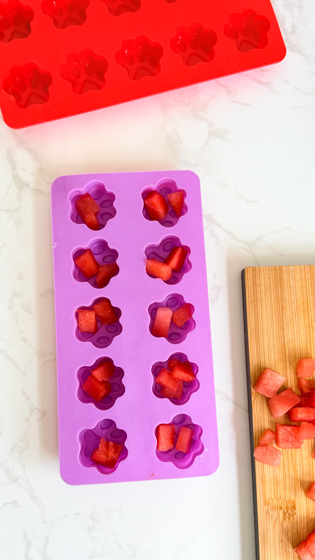 Purple silicone mould with paw-shaped compartments filled with pieces of watermelon, next to a chopping board with more watermelon and a red mould.