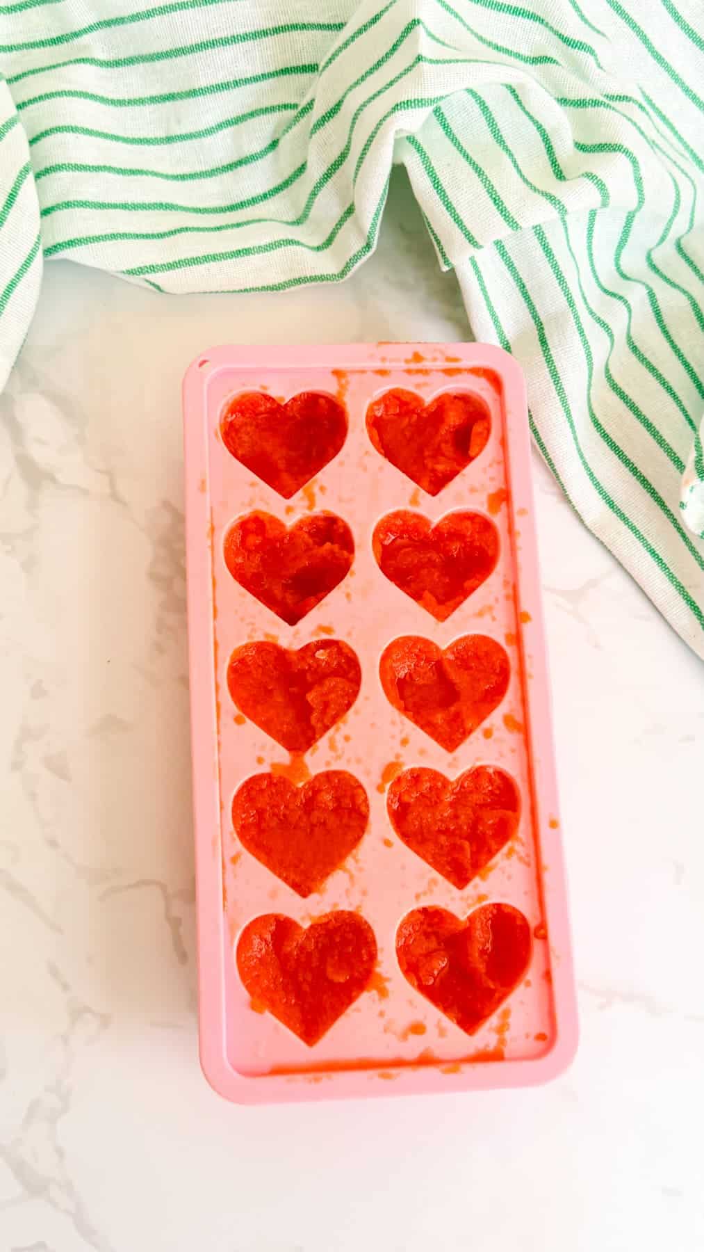 A pink silicone mould with ten heart-shaped compartments filled with a red mixture sits on a marble surface, with a green and white striped cloth nearby.