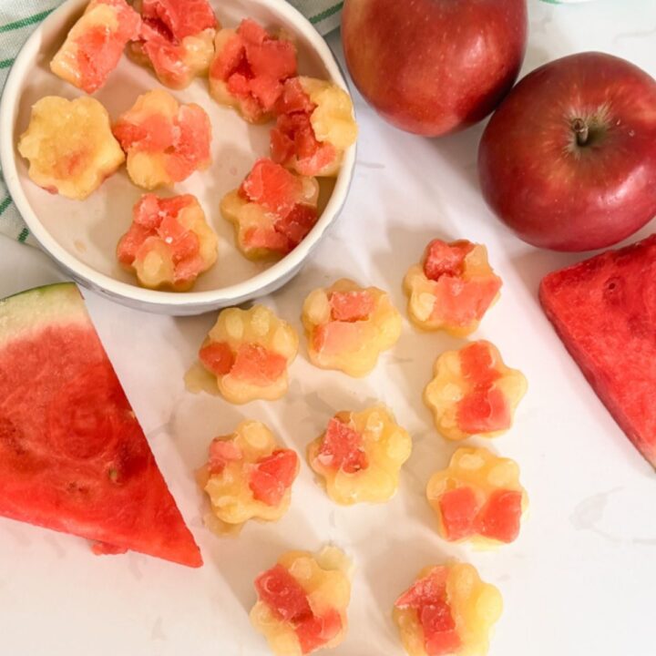 Flower-shaped iced lollies made with watermelon and apple, arranged on a white surface with fresh apples and watermelon slices nearby.