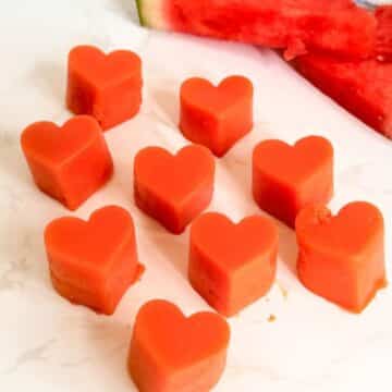 Nine heart-shaped pieces of watermelon are arranged on a white surface with two watermelon slices in the background.