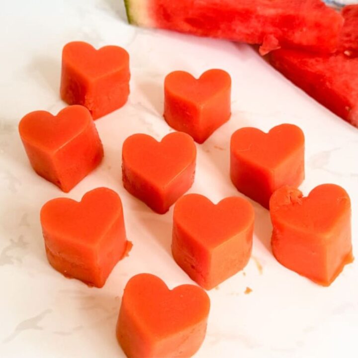 Nine heart-shaped pieces of watermelon arranged in rows on a white surface with watermelon slices in the background.
