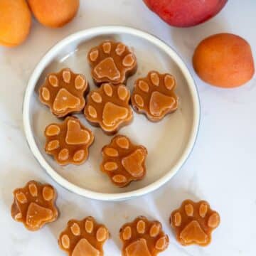 A white bowl filled with paw-shaped treats, surrounded by whole apricots and a peach on a white surface.