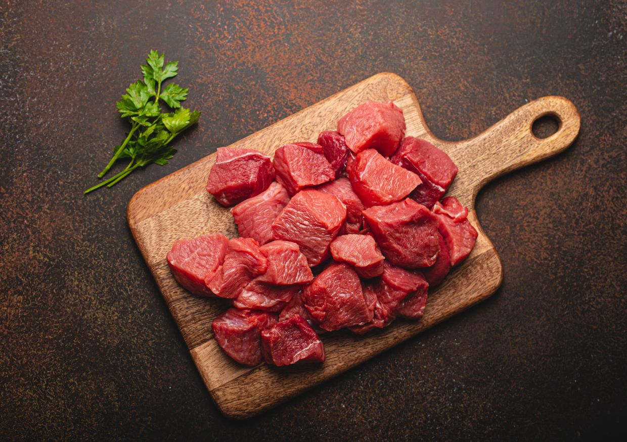 Chunks of raw beef, considered one of the best proteins for dogs, sit on a wooden chopping board with a small bunch of parsley on the side, placed on a dark textured surface.