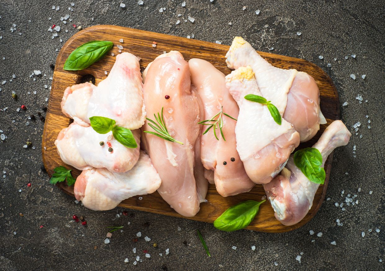 Raw chicken wings, drumsticks, and breasts—the best protein for dogs—are arranged on a wooden chopping board with basil leaves, rosemary, peppercorns, and coarse salt on a textured surface.