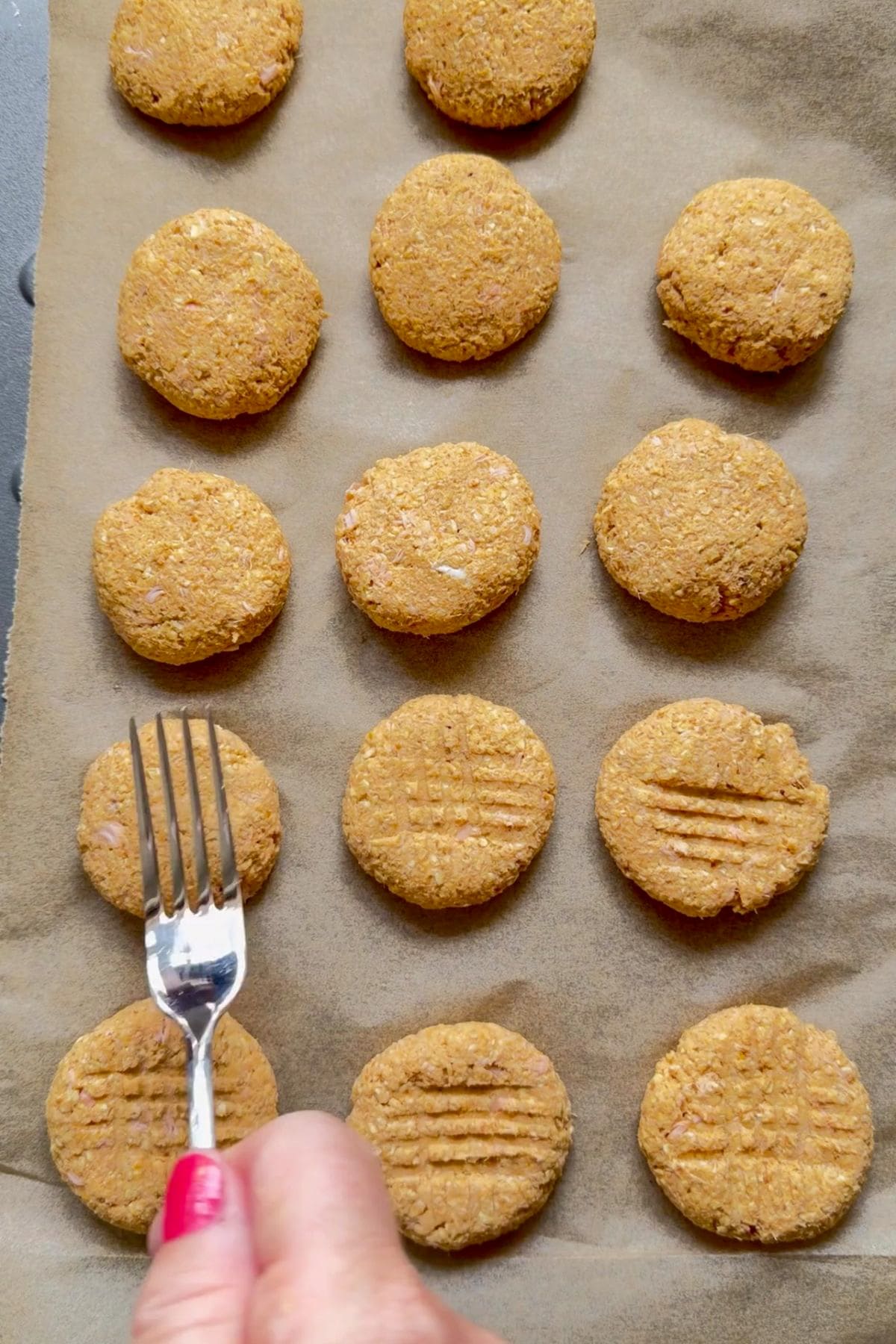 A hand presses a fork onto round, unbaked salmon pumpkin dog biscuits arranged in rows on baking parchment, creating a criss-cross pattern on some of them.
