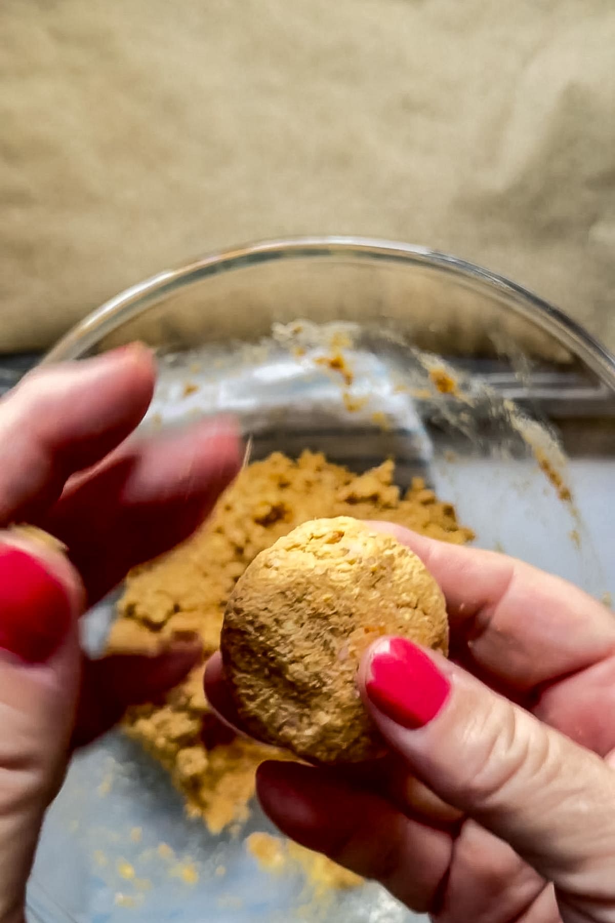 Hands with red nail varnish forming a round ball of crumbly salmon pumpkin dog biscuit mixture over a glass bowl filled with similar dough.