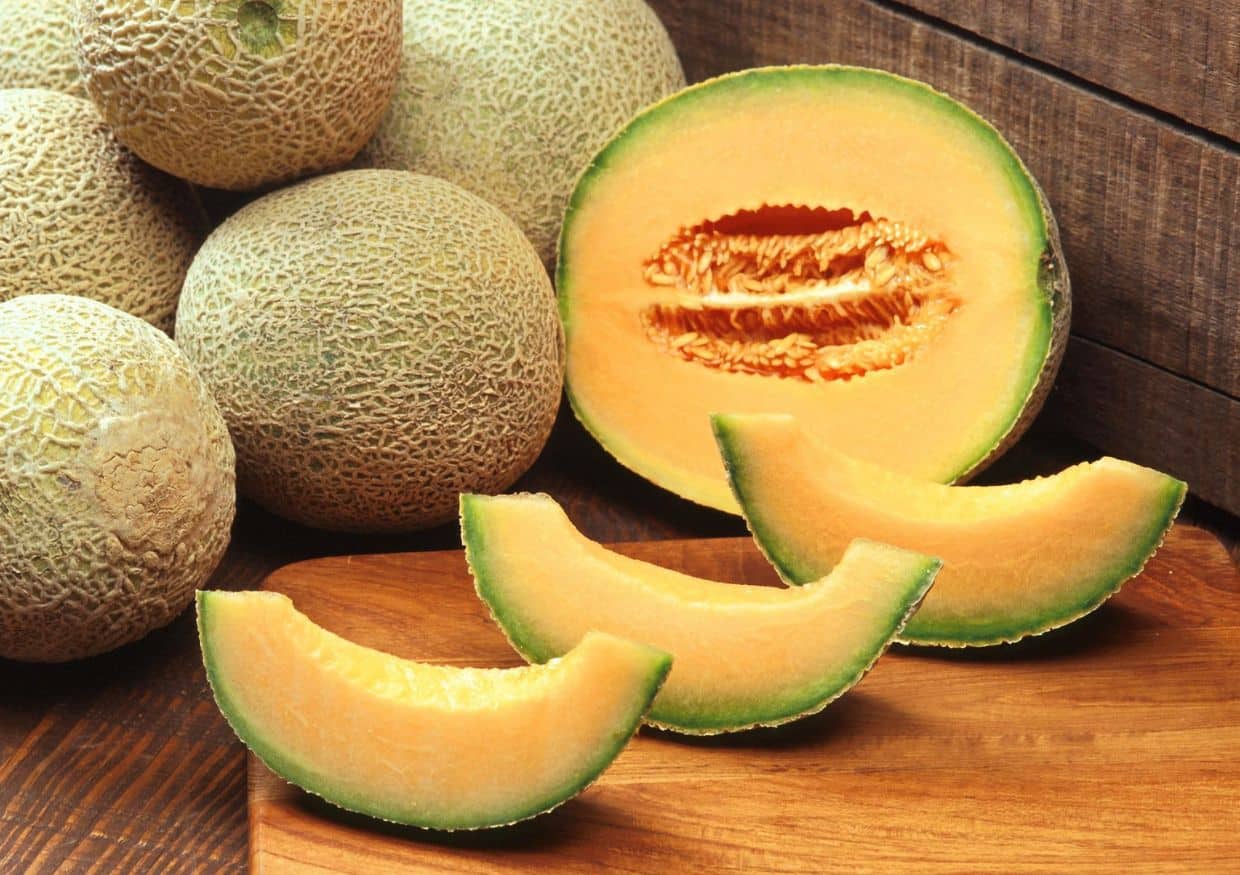 Several whole cantaloupes, one halved to show the orange flesh and seeds, with four cantaloupe wedges arranged on a wooden surface—prompting the question, can dogs eat cantaloupe?.