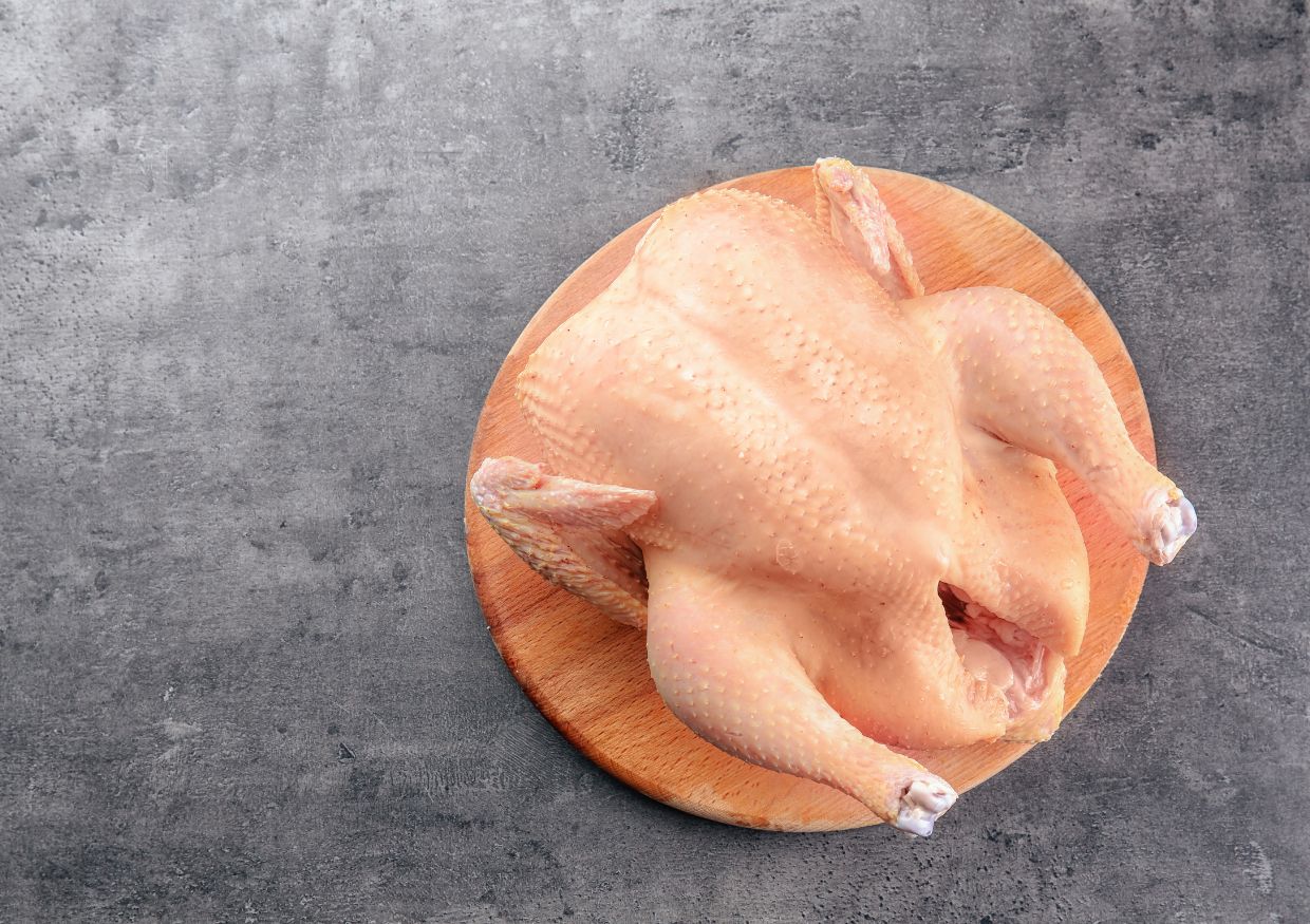 Raw whole chicken, considered one of the best proteins for dogs, is placed on a wooden chopping board, viewed from above on a grey textured surface.