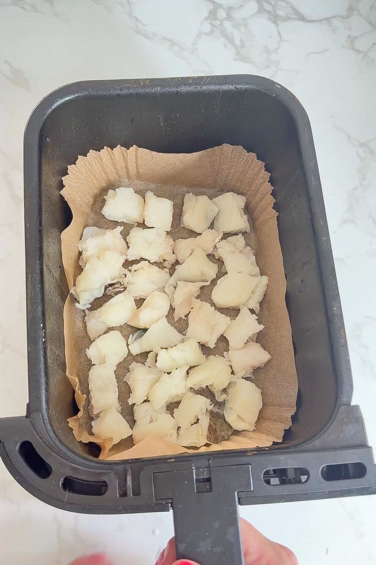 Chopped pieces of white fish, such as cod bites for dogs, placed on baking parchment in an air fryer basket, ready for cooking.