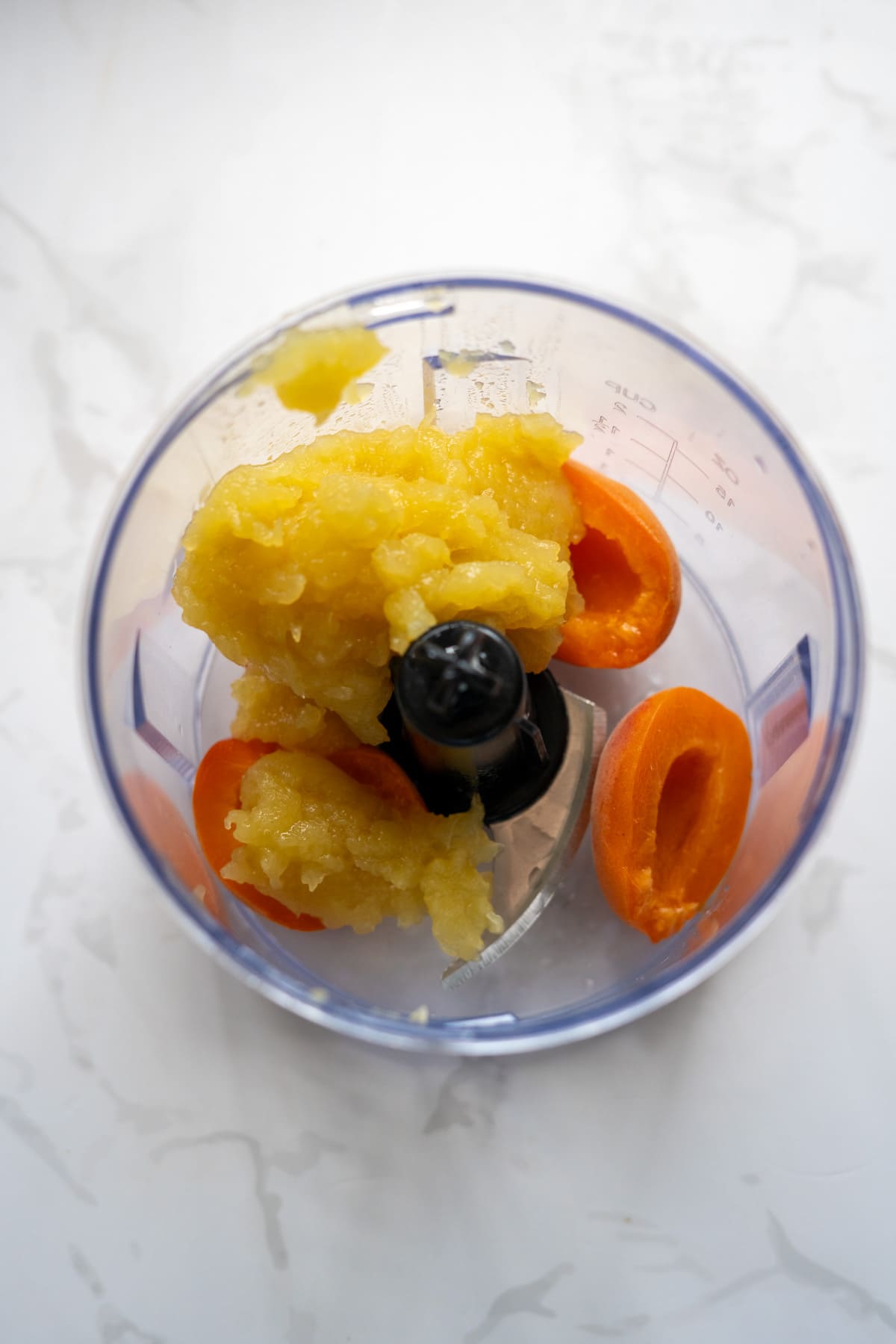 A food processor bowl containing chopped apricot halves and a mound of yellow fruit purée, viewed from above on a white surface.