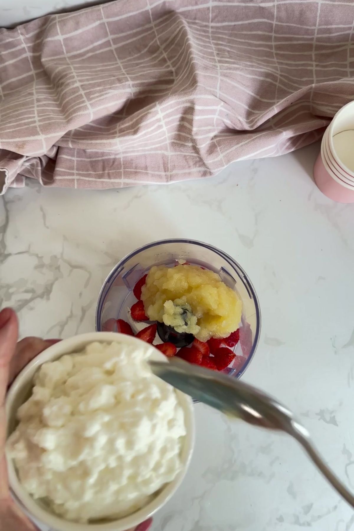 A hand holds a bowl of cottage cheese above a blender jug containing chopped strawberries, blueberries, and apple purée—perfect ingredients for making homemade apple strawberry dog ice cream—on a white worktop.