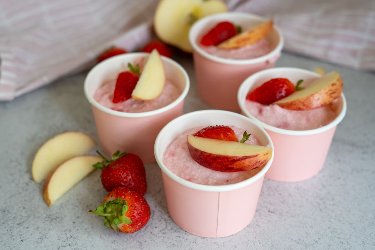Four pink cups filled with apple strawberry dog ice cream, each topped with a fresh strawberry and apple slice, sit on a light surface with extra strawberries and apple slices nearby.