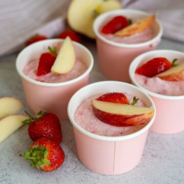 Four pink cups filled with a pink mousse-like dessert, each topped with a slice of peach and a strawberry half, surrounded by fresh strawberries and apple slices.