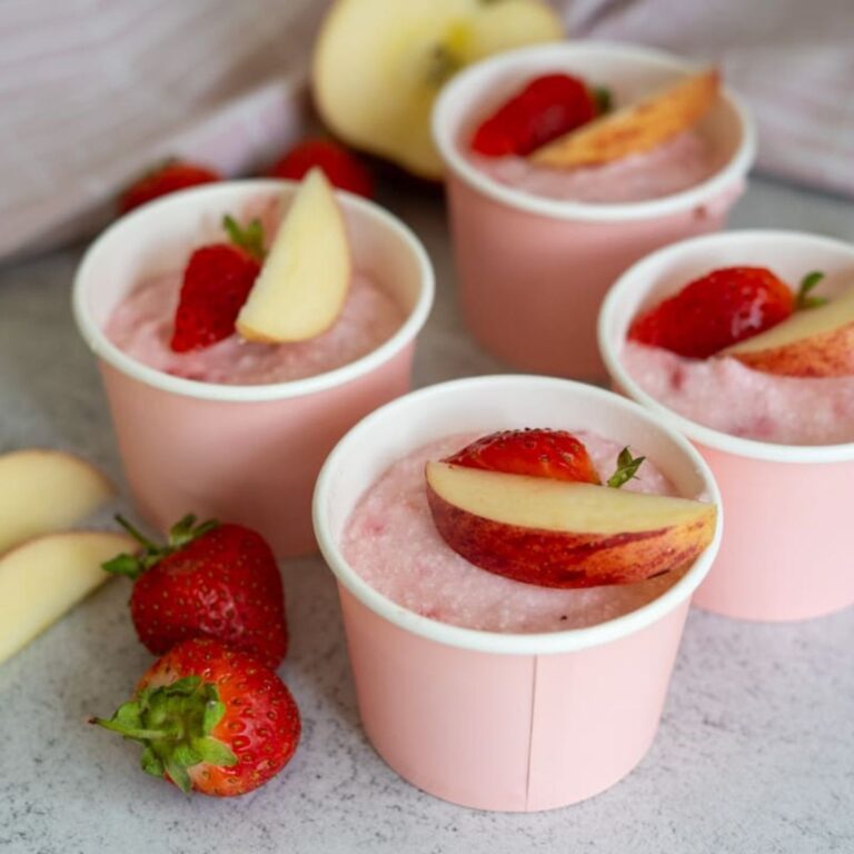 Four pink cups filled with a pink mousse-like dessert, each topped with a slice of peach and a strawberry half, surrounded by fresh strawberries and apple slices.