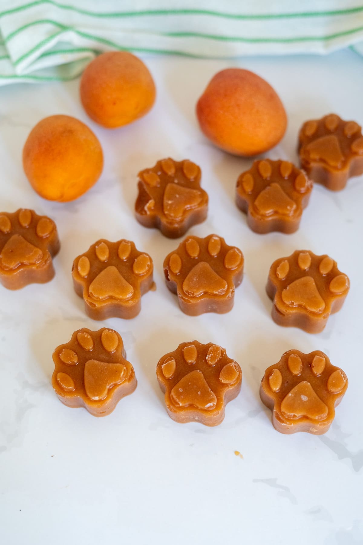 Paw-shaped treats are arranged on a white surface with three whole apricots and a striped cloth in the background.