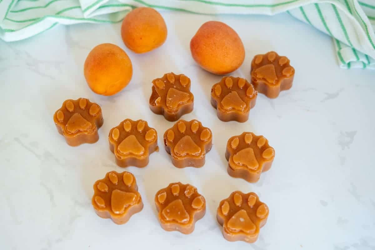 Ten paw-shaped brown treats are arranged on a white surface with three whole apricots and a green-striped cloth in the background.