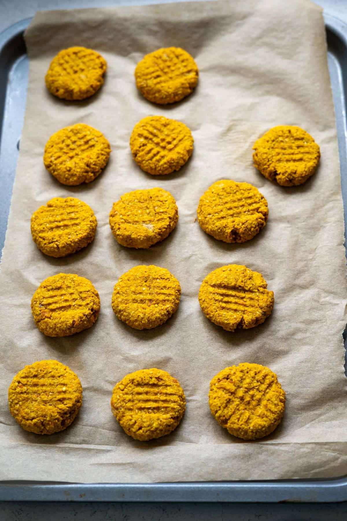 A baking tray lined with greaseproof paper holds 14 round, orange salmon pumpkin dog biscuits arranged in rows, each with a criss-cross fork pattern on top.