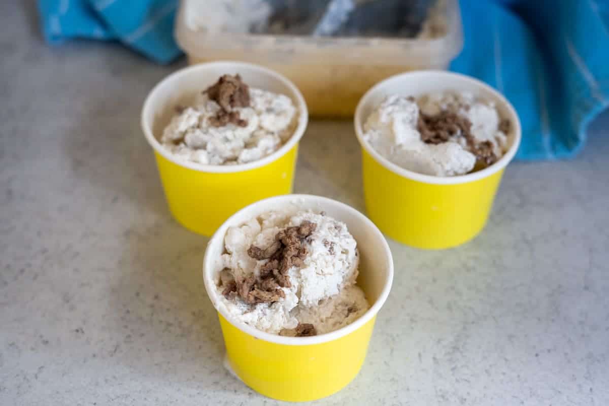 Three yellow cups filled with scoops of ice cream with chocolate chunks sit on a worktop, with an ice cream tub and blue towel in the background.