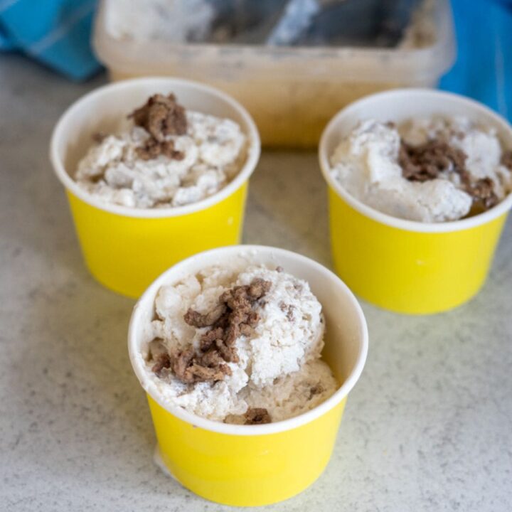 Three yellow cups filled with scoops of ice cream with a brown topping, placed on a light-coloured surface, with an ice cream tub in the background.