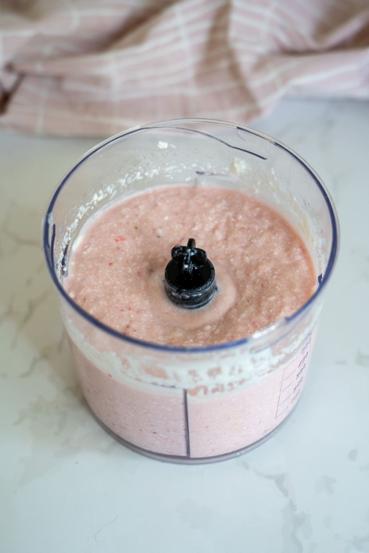 Clear plastic blender jug with a vibrant pink, apple and strawberry dog ice cream mixture inside, placed on a white marble worktop with a pink cloth in the background.