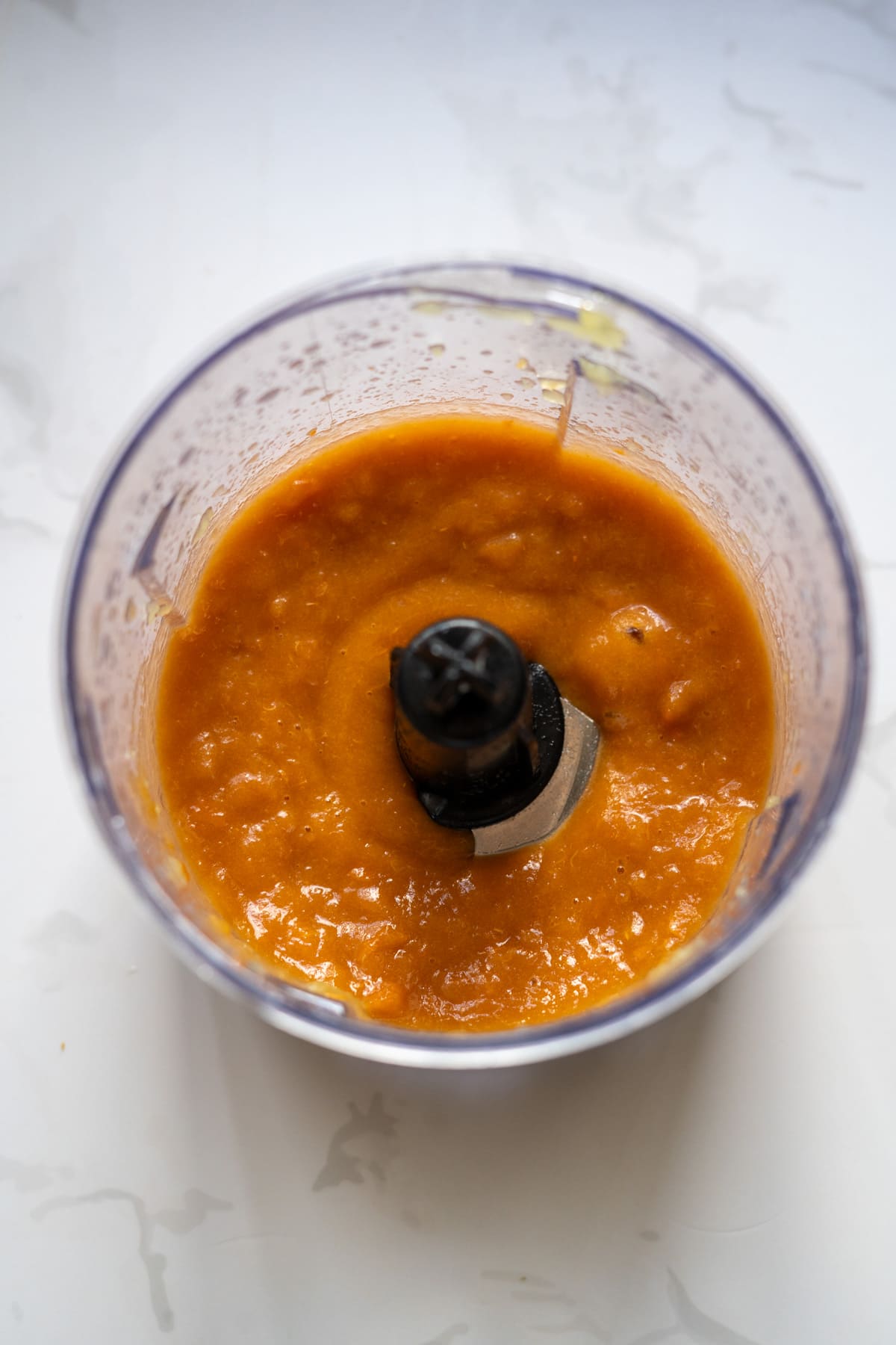 A food processor bowl containing a smooth orange purée, viewed from above on a white surface.