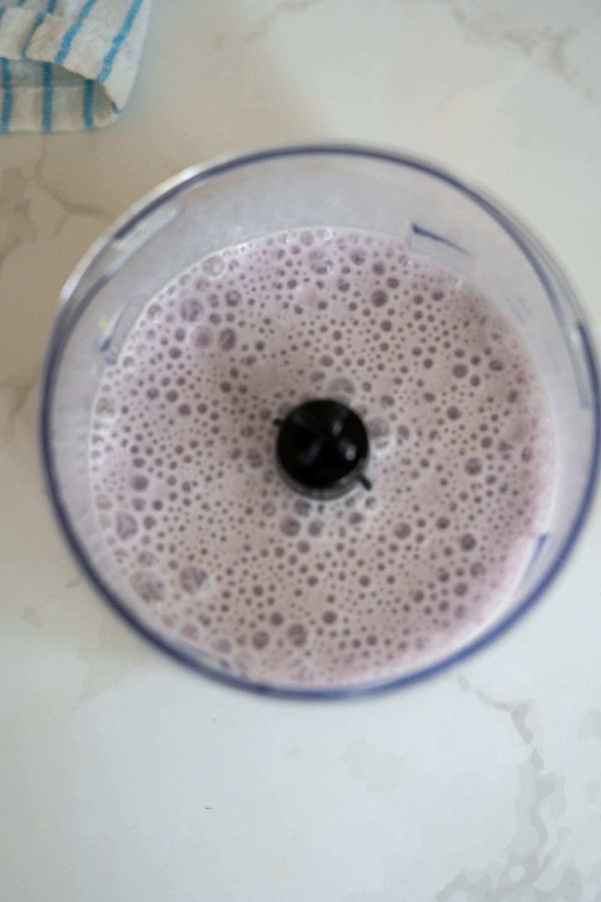 A blender jug filled with a light purple, frothy liquid sits on a white worktop next to a blue and white striped tea towel—perfect for whipping up homemade Frozen Goat Milk Dog Treats.